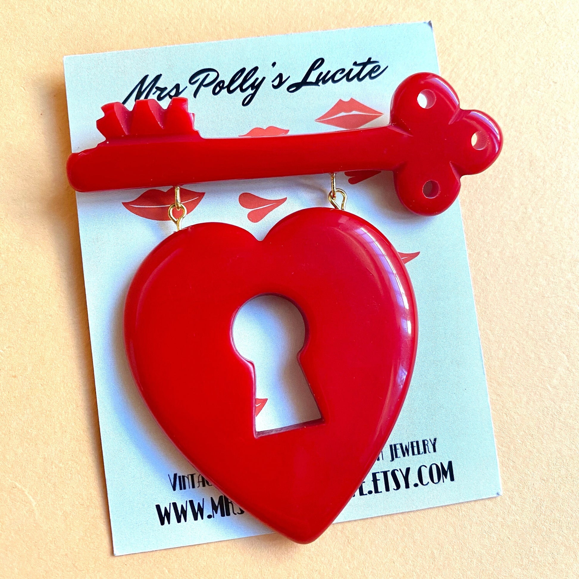 Red heart-shaped brooch with key hole hanging from a key, reproduction of the famous bakelite brooch from the 1940s placed on Mrs Polly&#39;s business card